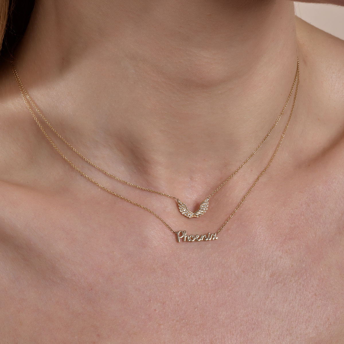 Gold diamond angel wings necklace layered with gold cursive phoenix necklace on model's neck, elegant layered jewelry