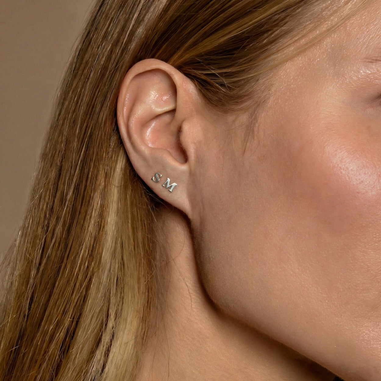 Close-up of a woman's ear wearing two small silver stud earrings shaped like the letters S and M.
