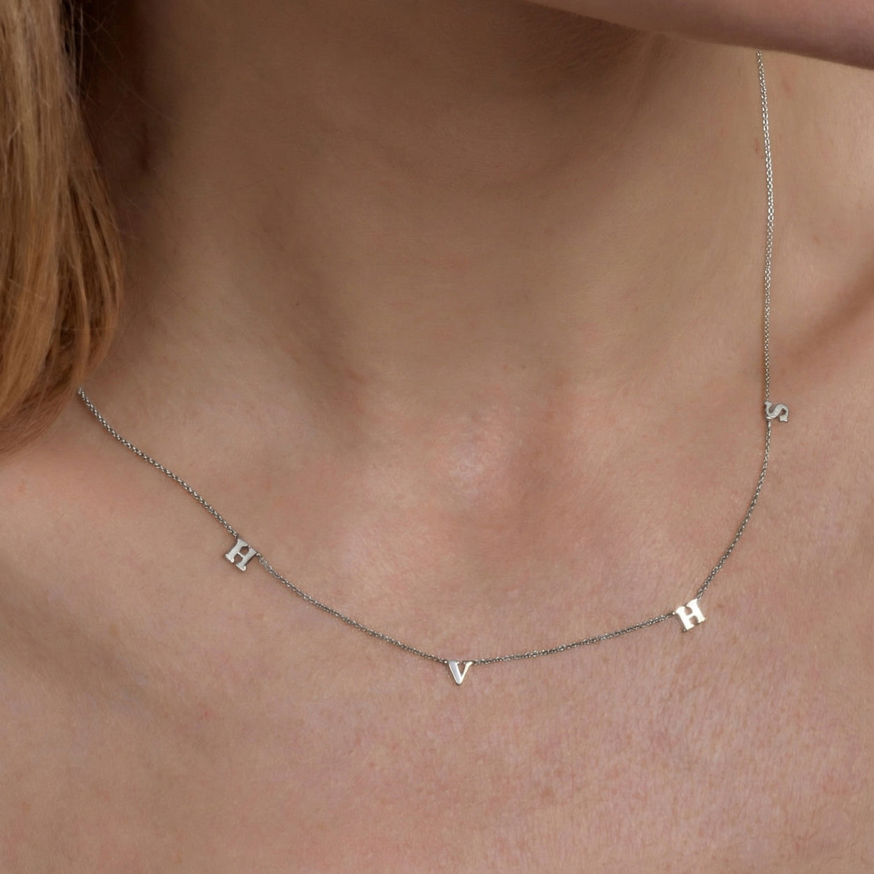 A silver chain necklace worn on the neck featuring spaced-out letter charms, including H, V, H, and S.