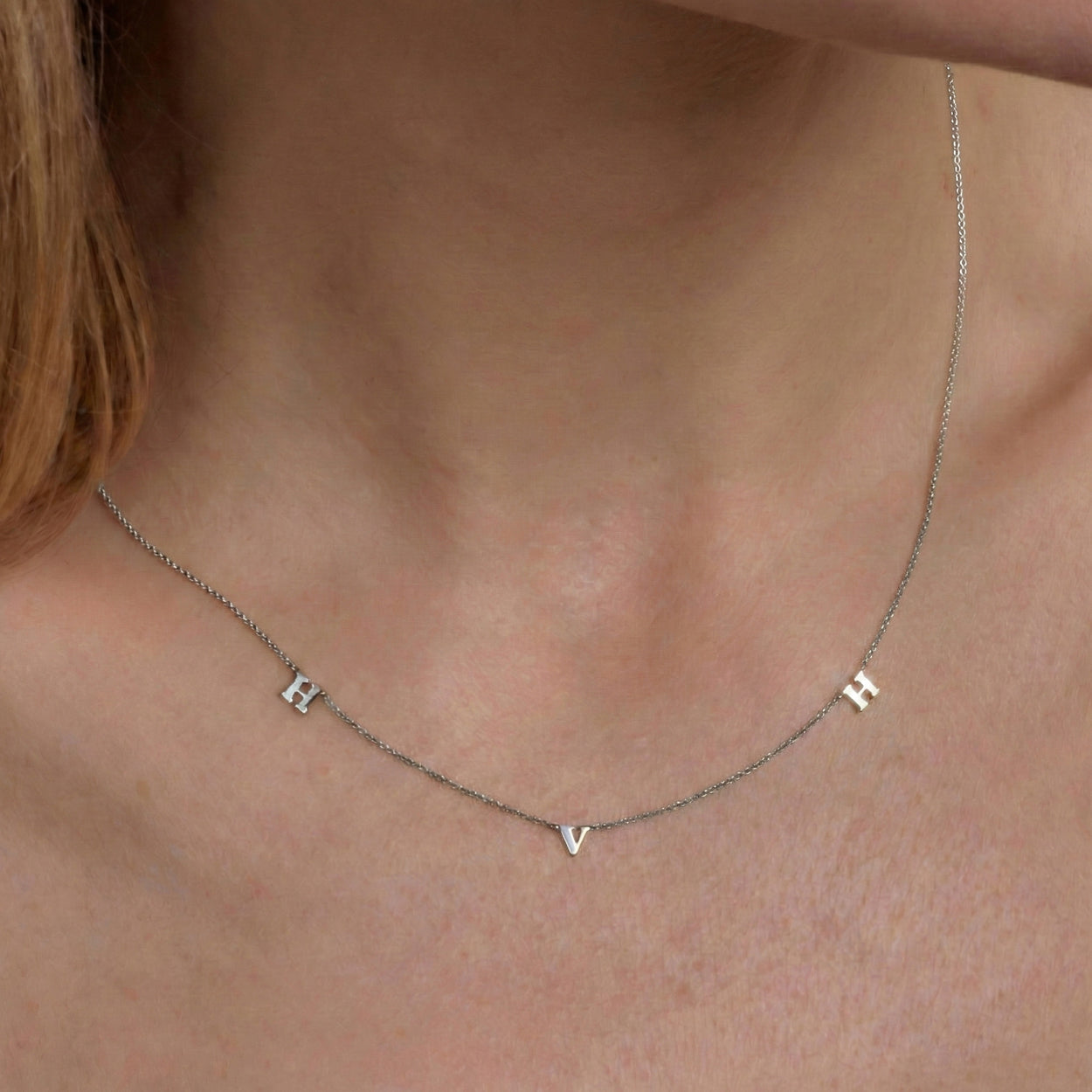 Close-up of a silver necklace with three spaced initial charms reading H, V, and H on a thin chain.